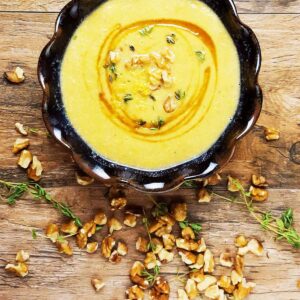 A bowl of yellow squash soup on a wooden table with walnuts and thyme scattered around it