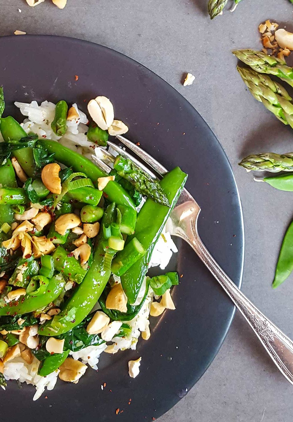 A dark plate with a stir fry of asparagus and snap peas, with nuts