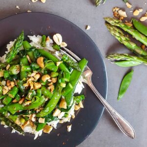 A dark plate with a stir fry of asparagus and snap peas, with nuts