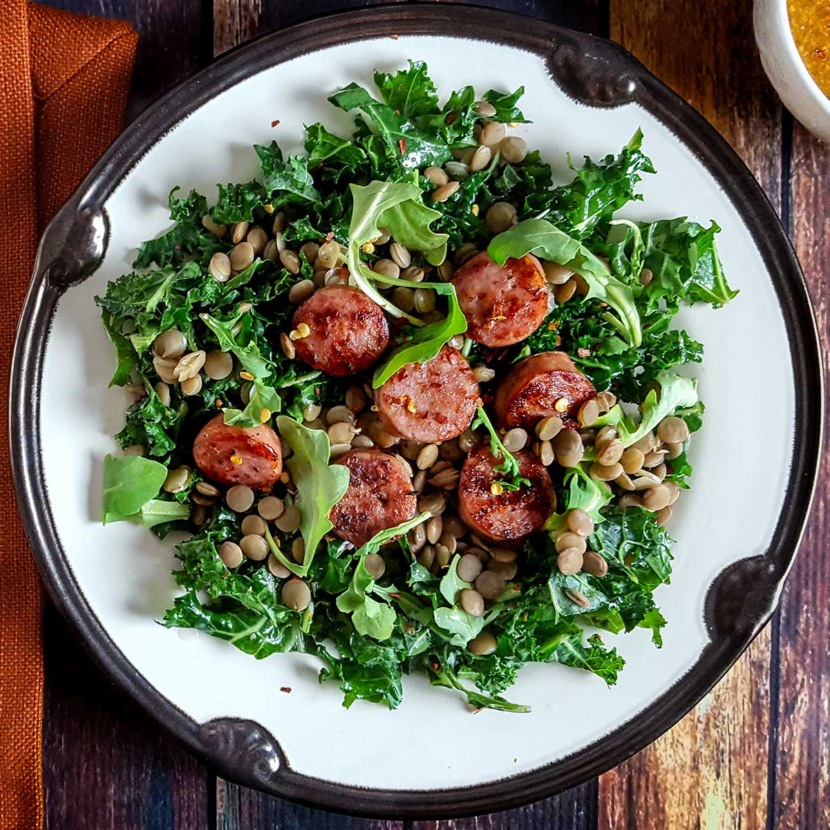 A plate of salad with kale, sausage, and lentils, on a wood table top