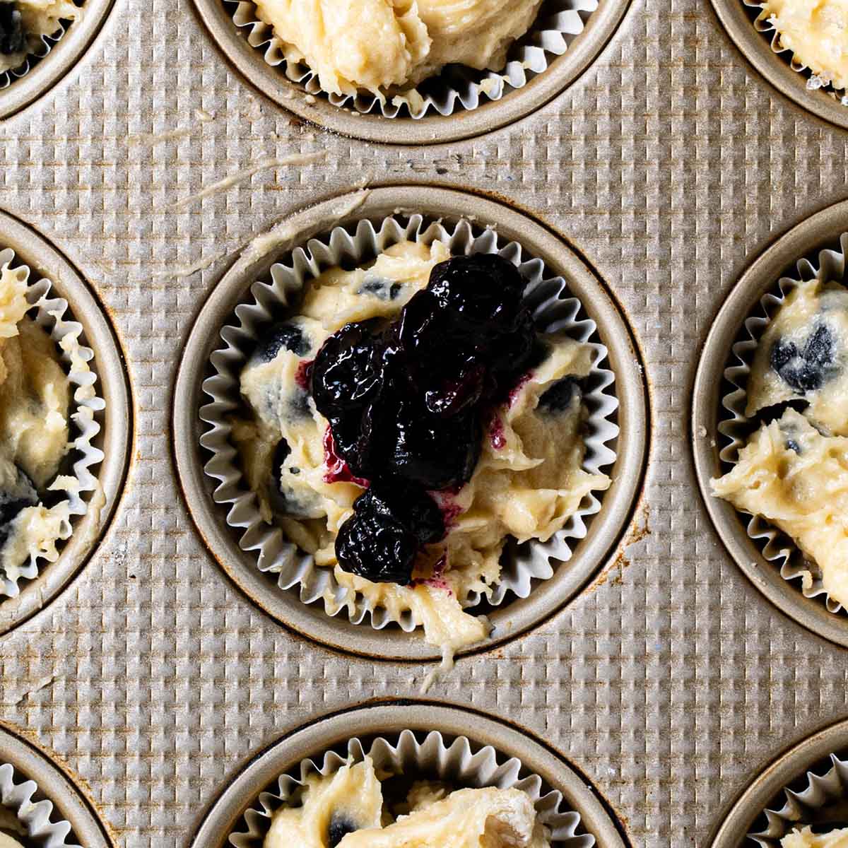 Blueberry muffin batter in a tin with a teaspoon of blueberry puree scooped on top.