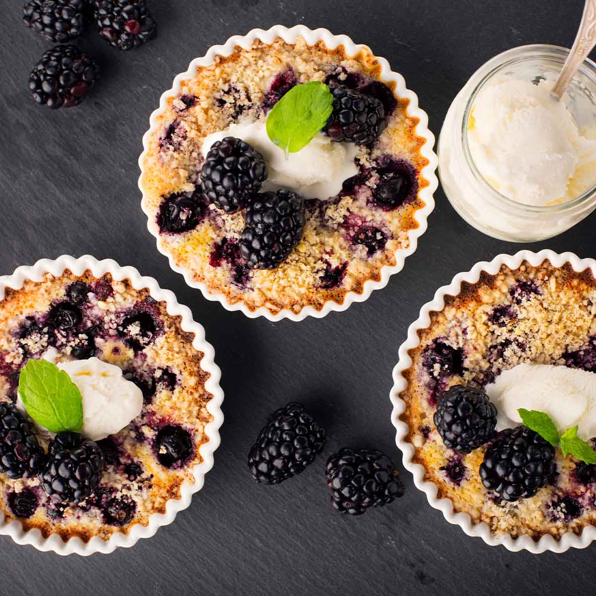 Blackberries are sweetened by being baked into mini cobblers, topped with whipped cream, on a dark background