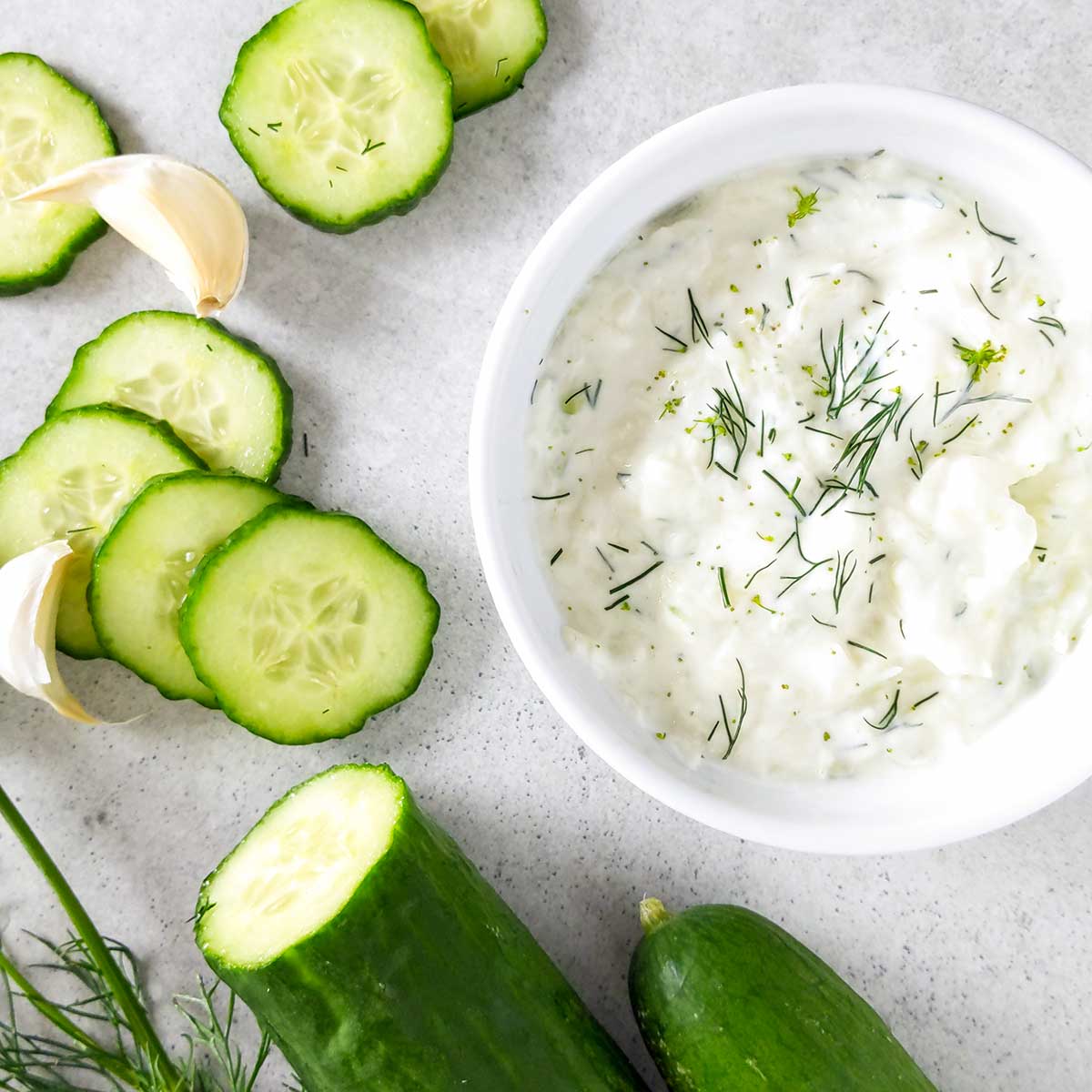 Bowl of tzatziki sauce surrounded by cucumbers and dill