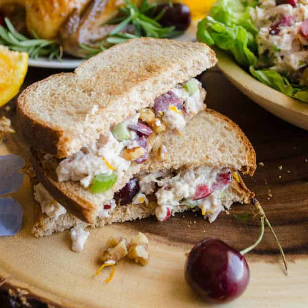 A chicken salad sandwich with cherries and candied walnuts, cut in half on a wood tray. Recipe from Goldilocks Kitchen.