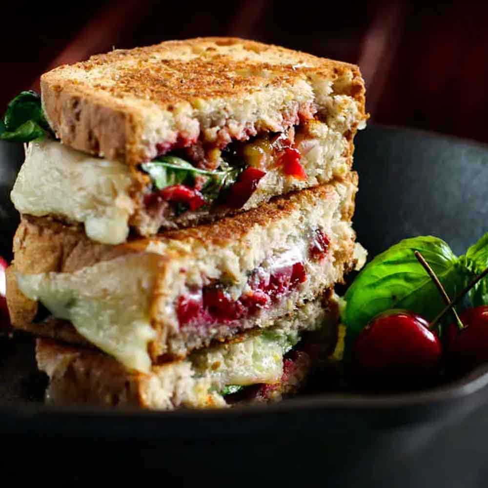 A grilled cheese sandwich with cherries and basil, cut in half and stacked on a black plate. Recipe from Cotter Crunch.