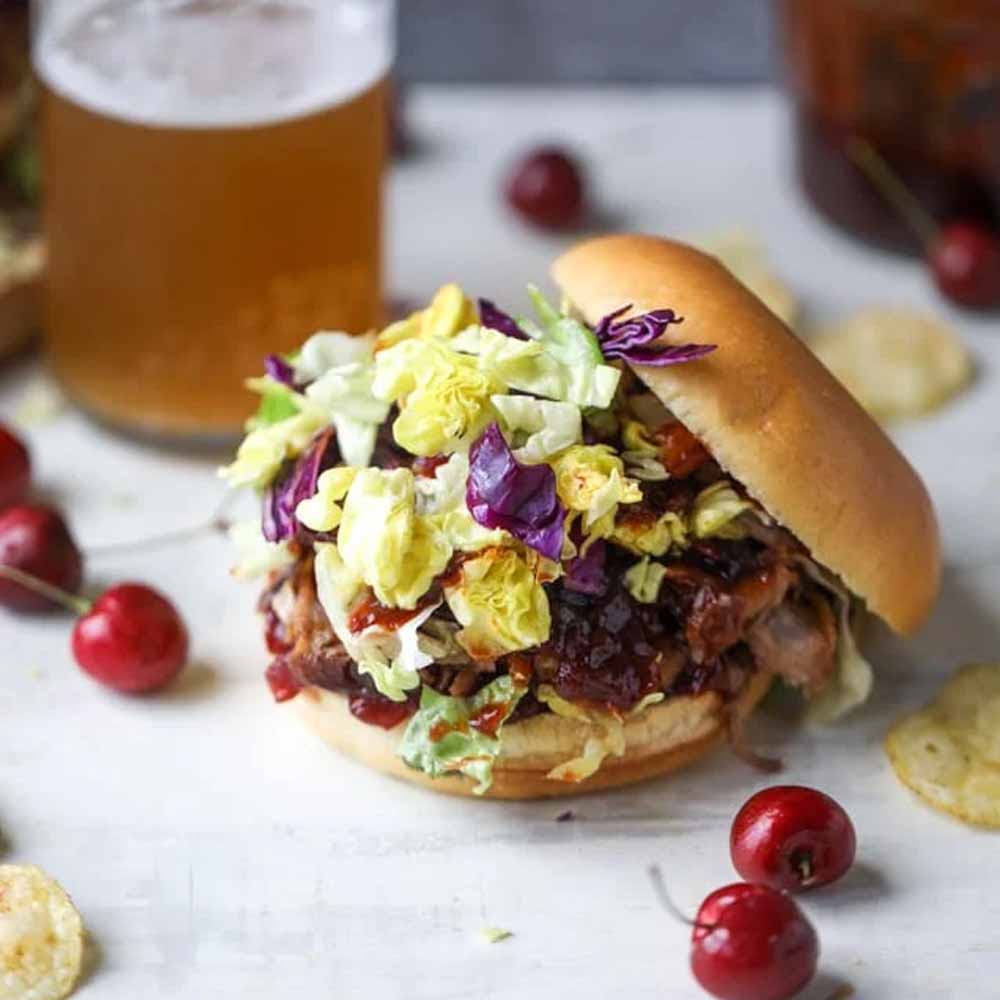 A pulled pork sandwich with fresh cherries on a table. Recipe from How Sweet Eats.