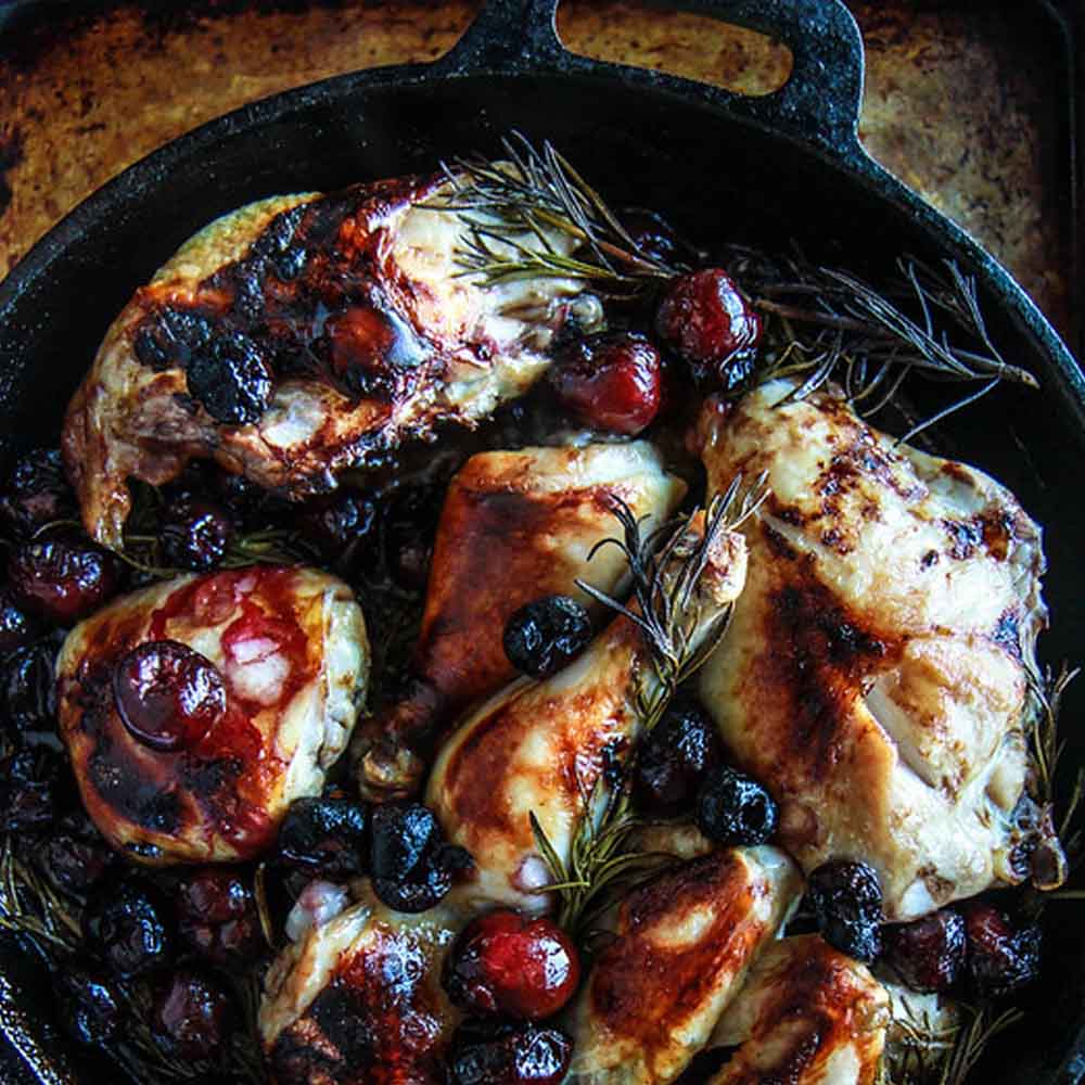 A cast iron pan with roasted chicken thighs, cherries, and rosemary, on a dark background. Recipe from Heather Christo.