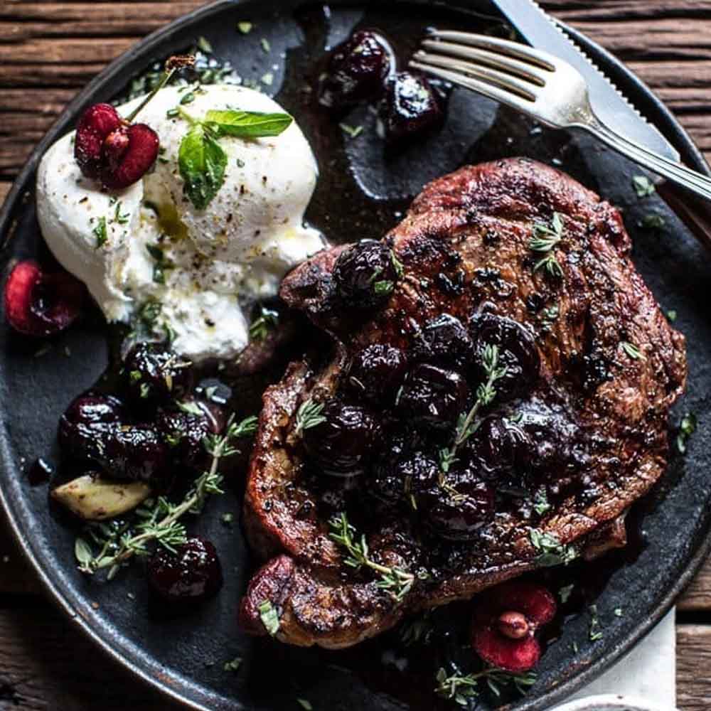 A steak on a dark plate topped with cherries with burrata cheese, on a wood table. Recipe from Half Baked Harvest