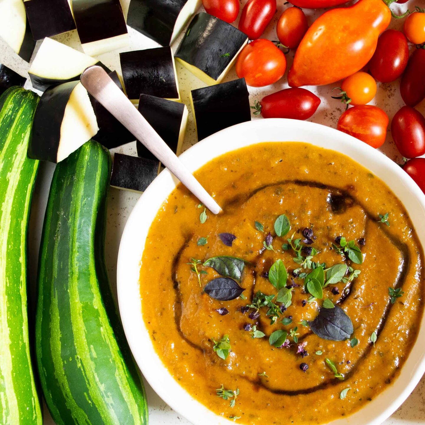 A bowl of Italian tomato zucchini soup on a table with eggplant, zucchini, and tomatoes.