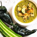 A bowl of zucchini poblano soup next to a green zucchini and poblano peppers on a table.