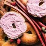 Homemade strawberry rhubarb cream cheese spread on a bagel, next to other bagels and fresh fruit.
