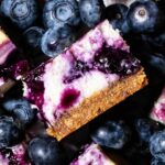 A blueberry cheesecake bar cut in half to show the cream cheese filling and graham cracker crust. Set on top of fresh blueberries.