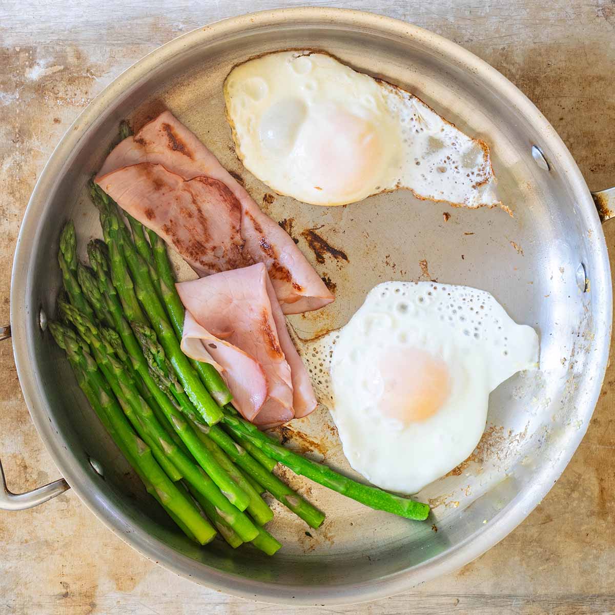 Eggs Benedict cooked in a pan, with the lid on to set the whites. A side of ham and asparagus are also in the pan.