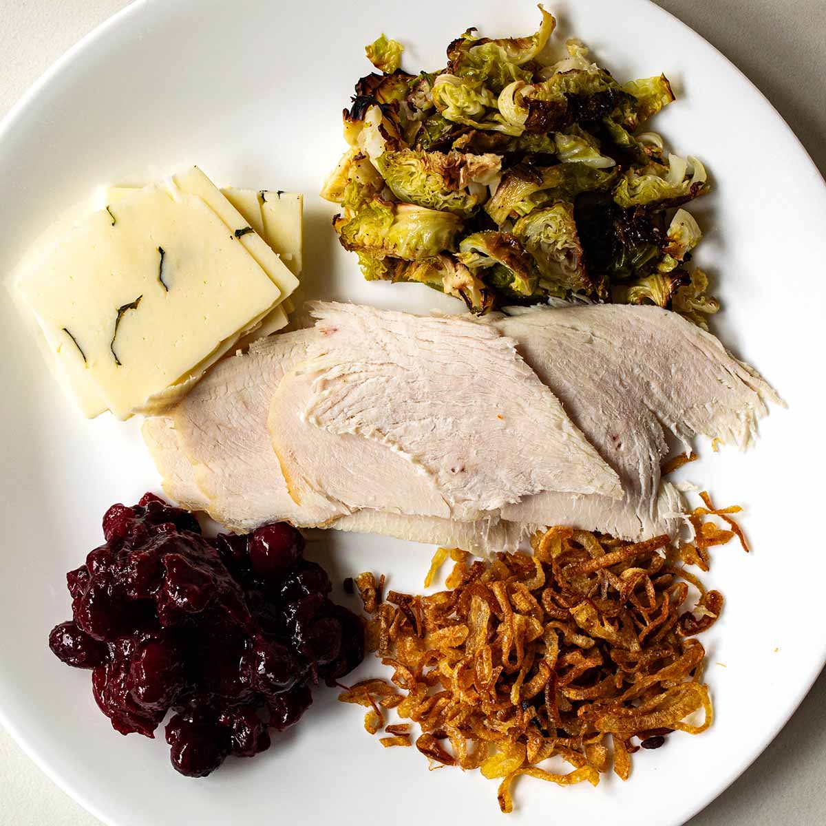A plate of leftovers from Thanksgiving, portioned out for a sandwich: Turkey breast, brussels sprouts, charcuterie cheese, cranberry sauce, and fried onions.